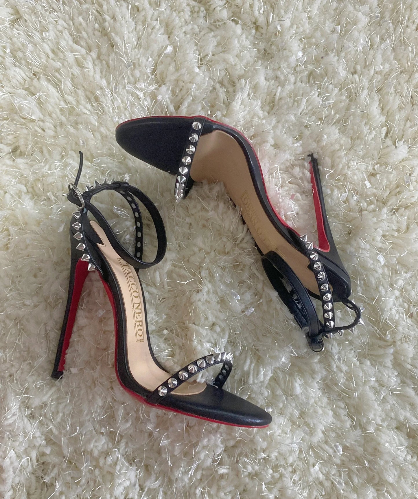 Black high heel sandals with silver studs and red soles on a textured cream carpet