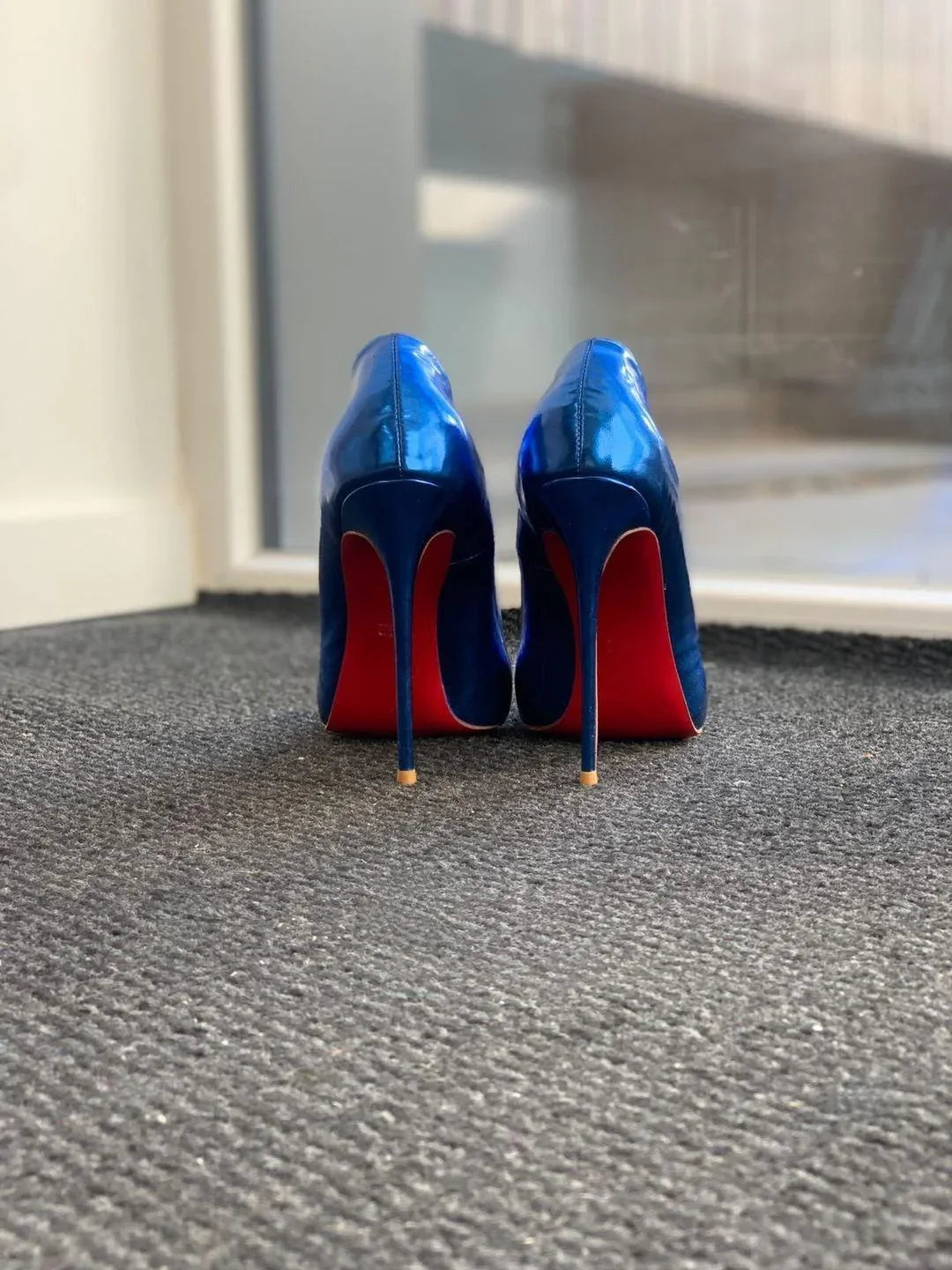 Blue patent leather high heels with red soles on textured gray carpet